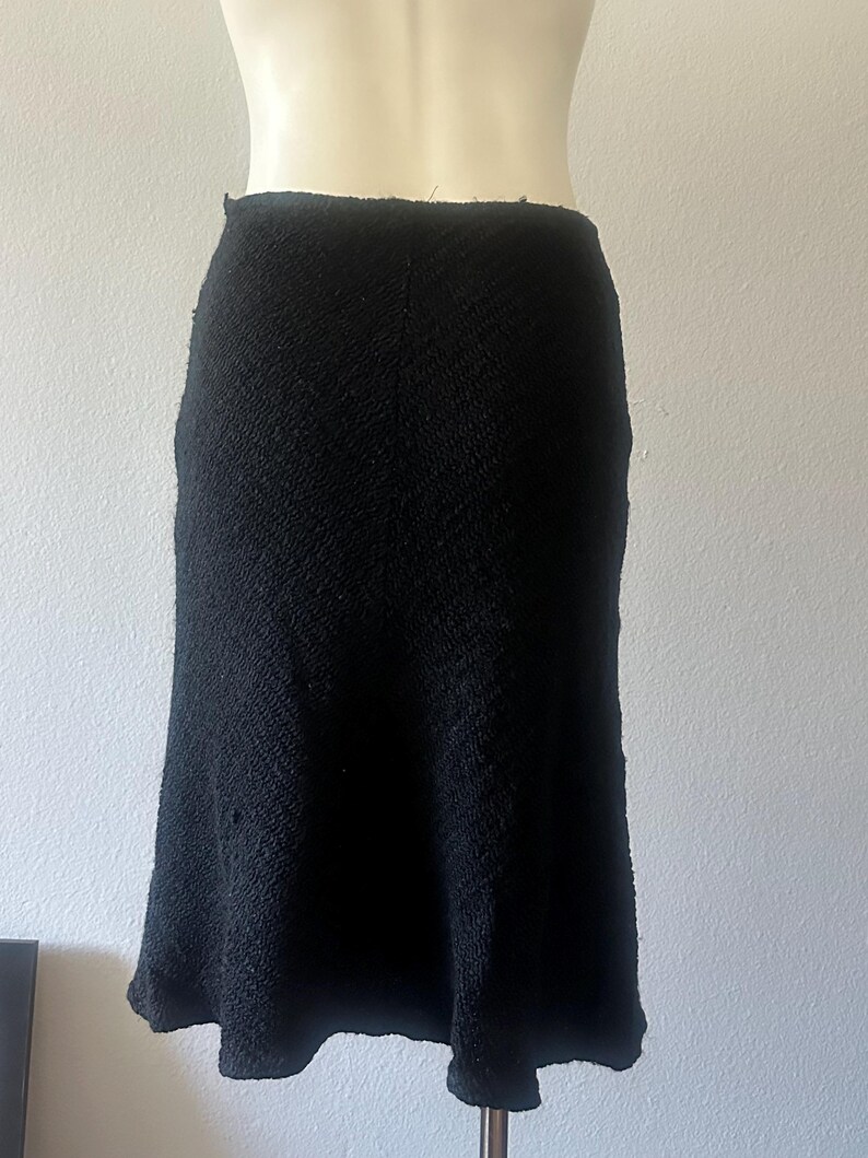 May include: A black, textured skirt with a slight A-line shape. The skirt appears to be made of a woven fabric, and it is displayed on a mannequin. The skirt's hemline falls below the knees.