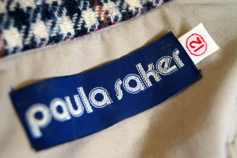 May include: A blue and white label with the text "Paula Saker" and a white label with the number "12" on a beige fabric background.