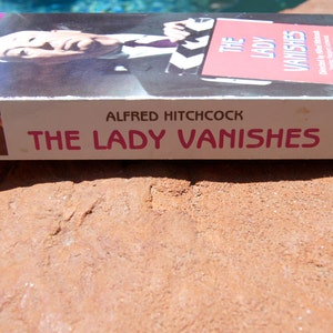 May include: A VHS tape of the Alfred Hitchcock film "The Lady Vanishes". The tape is white with pink and black text. The front of the tape features a black and white image of a man with a mustache.