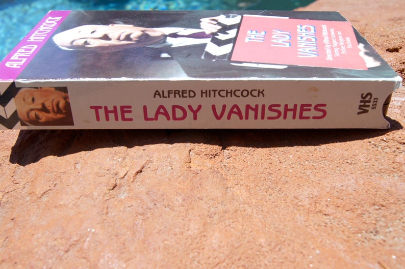 May include: A VHS tape of the Alfred Hitchcock film "The Lady Vanishes". The tape is white with pink and black text. The front of the tape features a black and white image of a man with a mustache.