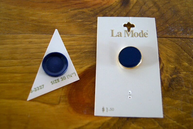 Op de afbeelding: Twee blauwe knopen met een gouden rand. De grotere knop bevindt zich op een witte kaart met de tekst "La Mode" en "&euro; 1,50". De kleinere knop bevindt zich op een witte driehoek met de tekst "Maat 30 (3/4")" en "4-3327".