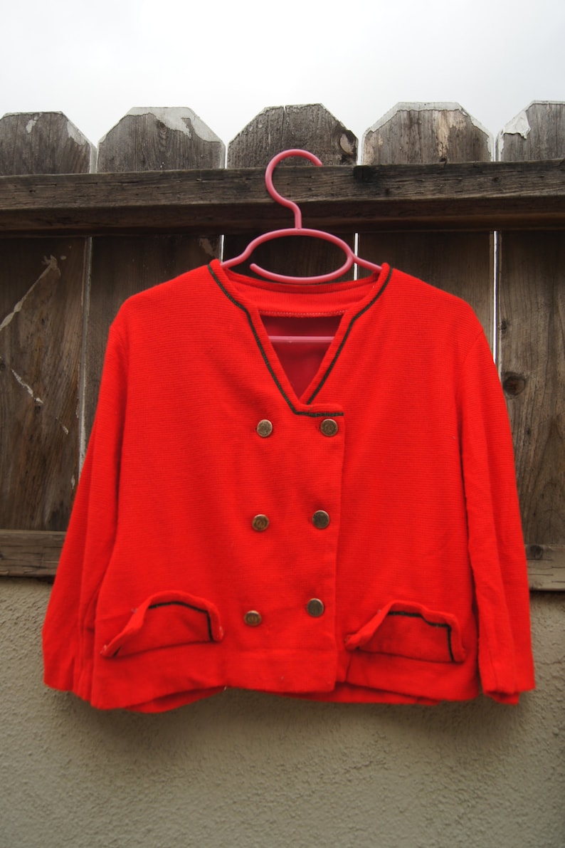 May include: A red double-breasted jacket with black trim and gold buttons. The jacket is hanging on a pink hanger.