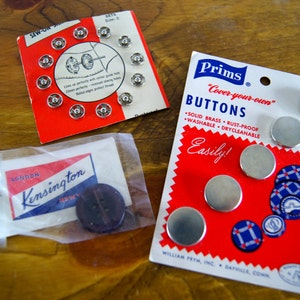 Op de afbeelding: Een close-up van twee pakjes naaiknopen. Het eerste pakje is rood en wit en bevat een verscheidenheid aan zilveren knopen. Het tweede pakje is rood, wit en blauw en bevat de tekst "Prims "Cover-your-own" Buttons" en "Easily!"  Het pakje bevat ook een verscheidenheid aan zilveren knopen.