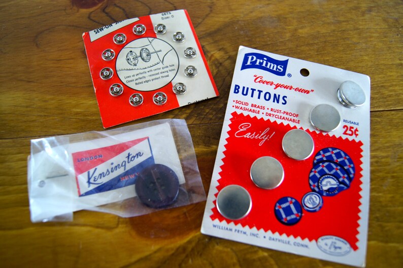 Op de afbeelding: Een close-up van twee pakjes naaiknopen. Het eerste pakje is rood en wit en bevat een verscheidenheid aan zilveren knopen. Het tweede pakje is rood, wit en blauw en bevat de tekst "Prims "Cover-your-own" Buttons" en "Easily!"  Het pakje bevat ook een verscheidenheid aan zilveren knopen.