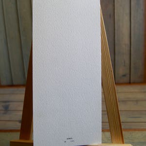 Op de afbeelding: Een witte kaart met een gestructureerd oppervlak wordt getoond op een houten ezel. De kaart heeft een klein zwart stipje en de tekst "253019" erop gedrukt.