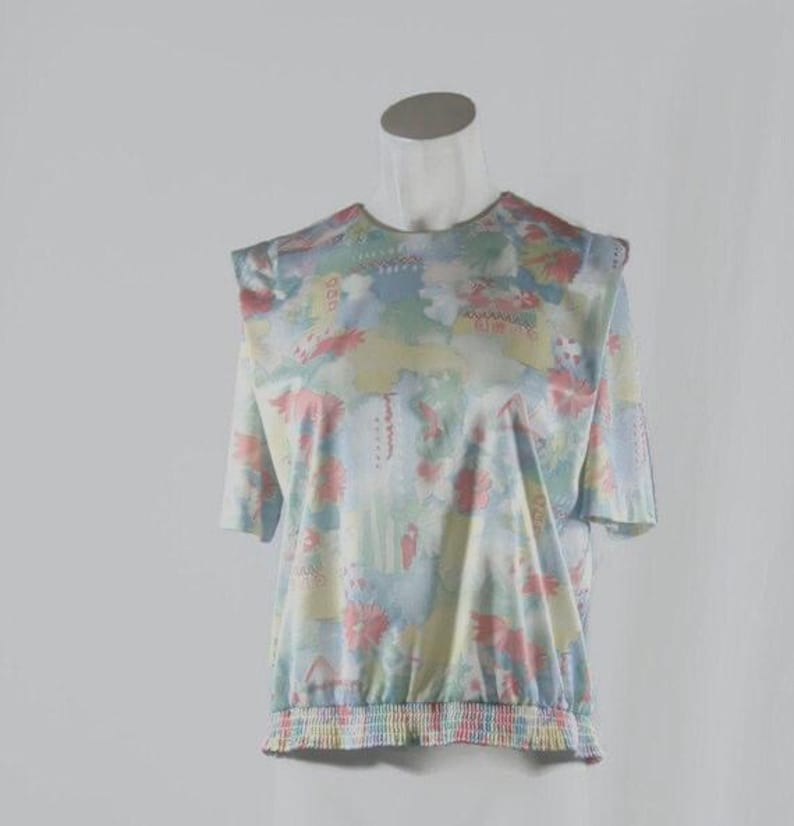 May include: A short-sleeved top with a floral pattern in pastel colors of pink, blue, yellow, and green. The top has a gathered elastic waist and a round neckline. The shoulders have a slight puff detail. The fabric appears to be lightweight.