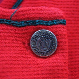 May include: Close-up of a red fabric with a green trim and a silver button with the text "SPLS X NOSIR" in a circle.