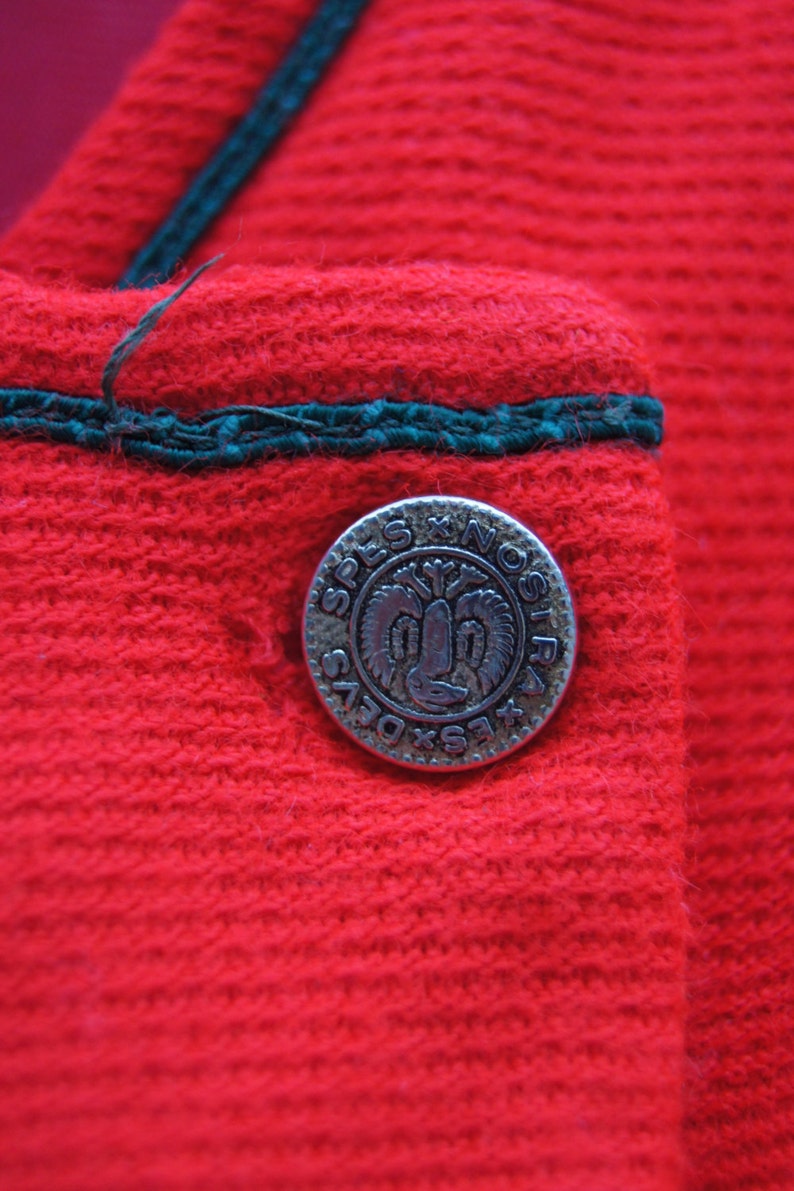 May include: Close-up of a red fabric with a green trim and a silver button with the text "SPLS X NOSIR" in a circle.