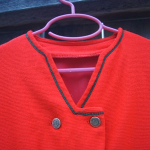 May include: Red cardigan with a V-neck and black trim. The cardigan has two gold buttons.