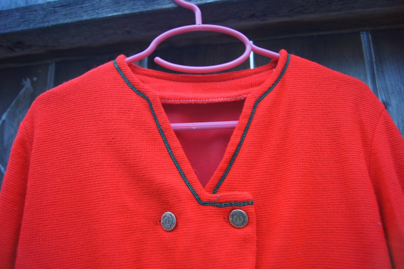 May include: Red cardigan with a V-neck and black trim. The cardigan has two gold buttons.