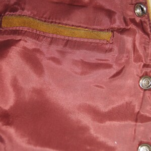 May include: Close-up of the inside of a brown leather jacket. The lining is a deep burgundy colour and has a small pocket on the left side. The jacket has metal poppers.