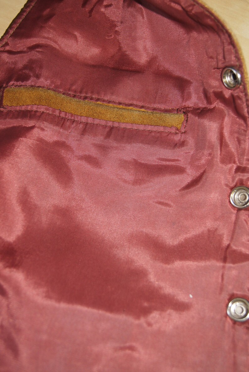 May include: Close-up of the inside of a brown leather jacket. The lining is a deep burgundy colour and has a small pocket on the left side. The jacket has metal poppers.