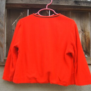 May include: A bright red long-sleeved sweater with a black and green trim around the neckline. The sweater is hanging on a pink hanger against a wooden fence.