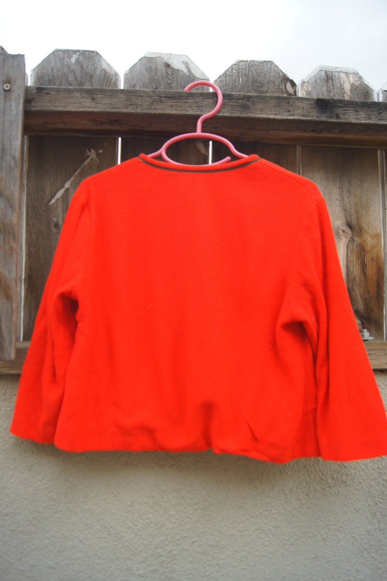 May include: A bright red long-sleeved sweater with a black and green trim around the neckline. The sweater is hanging on a pink hanger against a wooden fence.