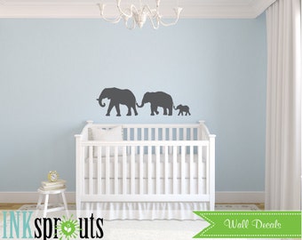 Könnte beinhalten: Ein Kinderzimmer mit einem weißen Babybett und einer blauen Wand. An der Wand befinden sich drei Elefanten-Silhouetten, eine große, eine mittlere und eine kleine. Die Elefanten gehen in einer Reihe, wobei der größte Elefant vorne und der kleinste Elefant hinten ist. Die Elefanten sind alle nach rechts gerichtet. Die Wand ist hellblau. Das Babybett ist weiß und hat eine weiße Matratze. Der Raum ist mit einem weißen Kronleuchter dekoriert.
