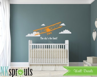 Könnte beinhalten: Orangefarbener Flugzeug-Wandaufkleber mit weißen Wolken und dem Text "The sky's the limit!" an einer blauen Wand. Der Aufkleber befindet sich über einem weißen Kinderbett mit einer weißen Matratze und einer weißen Decke.