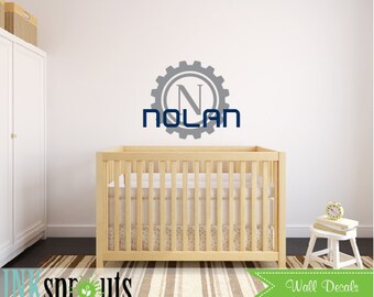 Könnte beinhalten: Ein graues und blaues Wandtattoo für ein Kinderzimmer mit einem Zahnrad-Design und dem Namen "NOLAN" in einem Kreis. Das Wandtattoo befindet sich an einer weißen Wand über einem Holzbettchen.
