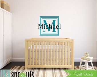 Könnte beinhalten: Blauer Monogramm-Wandaufkleber in Türkis mit dem Buchstaben M und dem Namen Michael in einem quadratischen Rahmen. Der Aufkleber befindet sich an einer weißen Wand über einem Holzbabybett.