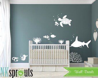Könnte beinhalten: Weiße Wandaufkleber mit einem Hai, einer Schildkröte, Fischen und Korallen. Die Aufkleber befinden sich an einer hellblauen Wand in einem Kinderzimmer mit einem weißen Kinderbett.