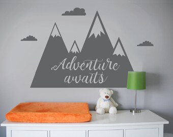 Könnte beinhalten: Ein grauer Wandaufkleber mit einer Bergkette und dem Text "Adventure awaits" an einer grauen Wand. Ein weißer Teddybär sitzt auf einer weißen Kommode mit einer orangefarbenen Wickelauflage.