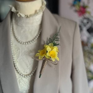 May include: A yellow daffodil buttonhole with a brown twine bow and green foliage.