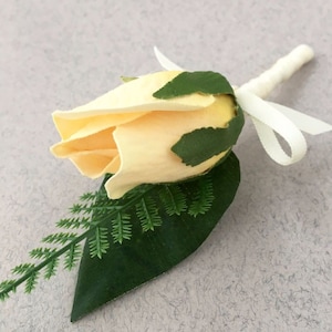May include: A cream-coloured rose buttonhole with green leaves and a white ribbon. The artificial flower has a realistic appearance, with detailed petals and foliage. The stem is wrapped in a cream-coloured fabric.