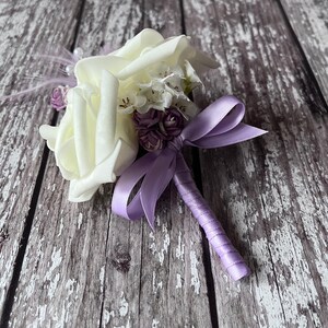以下が含まれることがあります： 白いバラのブートニアに、ラベンダーのリボンと小さな紫色の花束が添えられています