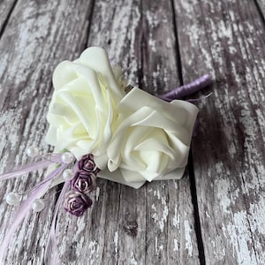 以下が含まれることがあります： 2つの大きな白いバラと小さな紫色の花が付いた白い造花のコサージュ。コサージュは紫のリボンで留められています。