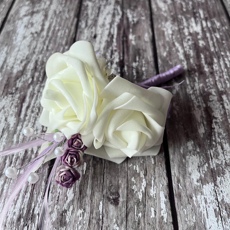 以下が含まれることがあります： 2つの大きな白いバラと小さな紫色の花が付いた白い造花のコサージュ。コサージュは紫のリボンで留められています。
