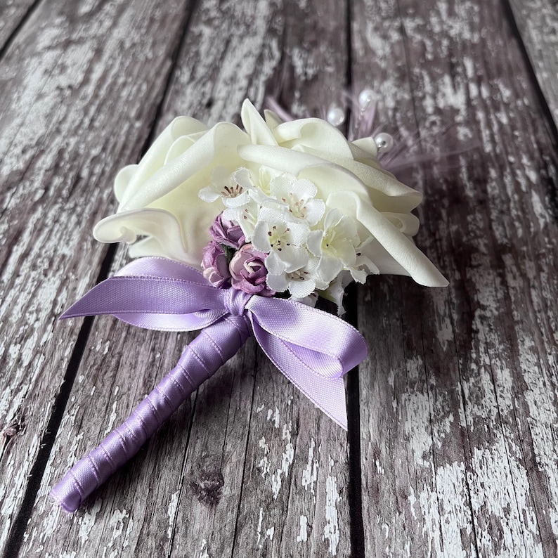 以下が含まれることがあります： 白い花と紫の花のブートニアで、サテンのリボンが付いています。花は房状にまとめられ、紫のリボンで結ばれています。