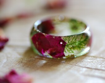 May include: A clear resin ring with embedded dried rose petals and green moss. The ring is a wide band and has a smooth, polished finish.