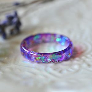 May include: A close-up of a purple resin ring with iridescent glitter and flecks. The ring is set against a blurred background with lavender sprigs. The ring's colors include purple, blue, and green.