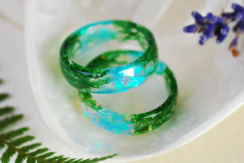 Op de afbeelding: Twee groene en blauwe harsringen met ingebed groen gebladerte en gouden glitter. De ringen zijn op elkaar gestapeld.