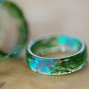 Op de afbeelding: Een ring van helder hars met groen mos en iriserende glitter erin ingebed. De ring is dik en heeft een afgeronde vorm.