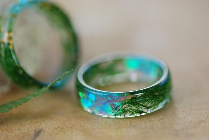 Op de afbeelding: Een ring van helder hars met groen mos en iriserende glitter erin ingebed. De ring is dik en heeft een afgeronde vorm.