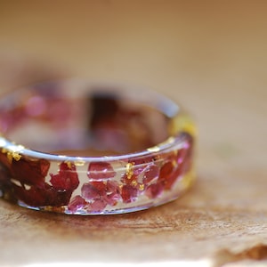 May include: A clear resin ring with embedded red and gold flakes. The ring is thick and has a rounded shape.