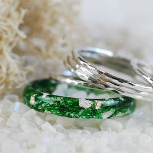 May include: Close-up of three rings. One ring is green with embedded moss and silver flakes. Two silver rings with a hammered texture are stacked behind the green ring. The rings are on a bed of white stones.