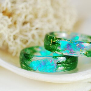 Op de afbeelding: Twee groene harsringen met ingebedde iriserende blauwe en groene glitter en gouden vlokken. De ringen zijn op elkaar gestapeld en liggen op een witte schelp.