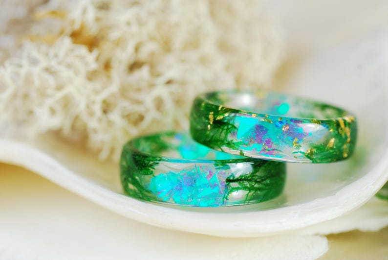 Op de afbeelding: Twee groene harsringen met ingebedde iriserende blauwe en groene glitter en gouden vlokken. De ringen zijn op elkaar gestapeld en liggen op een witte schelp.