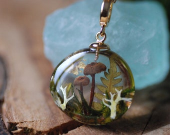 May include: A gold pendant with a clear resin dome containing a miniature forest scene. The scene includes small mushrooms, ferns, and moss.