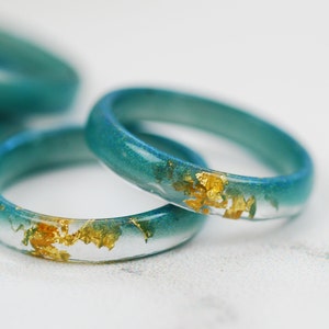 May include: Three teal blue resin rings with gold flakes embedded in the resin. The rings are stacked on top of each other.