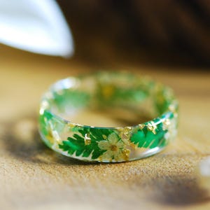 May include: A clear resin ring with embedded green leaves, small white flowers, and gold flakes. The ring is set against a blurred background with a white flower and other floral elements.