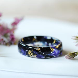 Peut inclure: Une bague en résine noire ornée de pétales de fleurs bleues et de feuilles d'or. La bague est posée sur une surface blanche, avec des fleurs roses en arrière-plan. Un bijou unique.