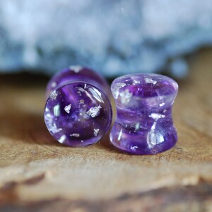 Puede incluir: Dos expansores de oreja de color morado y plateado con un acabado brillante. Los expansores est&aacute;n hechos de resina y tienen una apariencia clara y transl&uacute;cida.