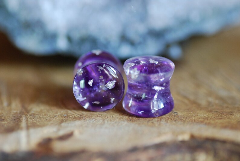 Puede incluir: Dos expansores de oreja de color morado y plateado con un acabado brillante. Los expansores est&aacute;n hechos de resina y tienen una apariencia clara y transl&uacute;cida.