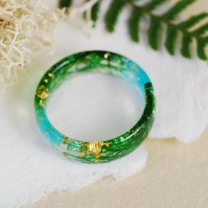 Op de afbeelding: Een groene en blauwe harsring met gouden vlokken ingebed in de hars. De ring ligt op een wit oppervlak.