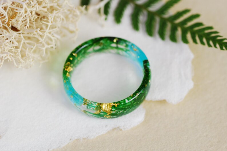 Op de afbeelding: Een groene en blauwe harsring met gouden vlokken ingebed in de hars. De ring ligt op een wit oppervlak.