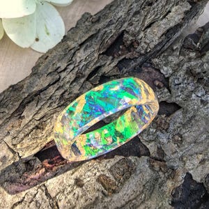 Pode incluir: Um anel de resina transparente com fragmentos iridescentes verdes e azuis incrustados, juntamente com flocos de folha de ouro. A banda tem um acabamento liso e polido, exibindo um efeito reflexivo multicolorido vibrante no material.