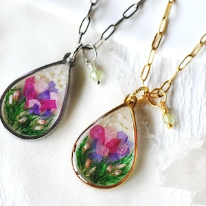May include: Two teardrop-shaped pendant necklaces. One has a silver-colored chain and frame, the other a gold-colored chain and frame. Both pendants contain dried flowers in shades of pink, purple, and green. Each necklace has a small green charm.
