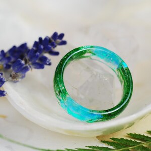 Op de afbeelding: Een groene en blauwe harsring met ingebedde botanische elementen. De ring ligt op een wit oppervlak met lavendelbloemen en groene bladeren.